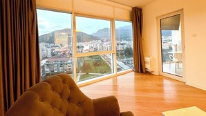 Ein geräumiges Zimmer mit großen Fenstern und Blick auf die Stadt, ein bequemer Sessel und eine moderne Deckenleuchte.