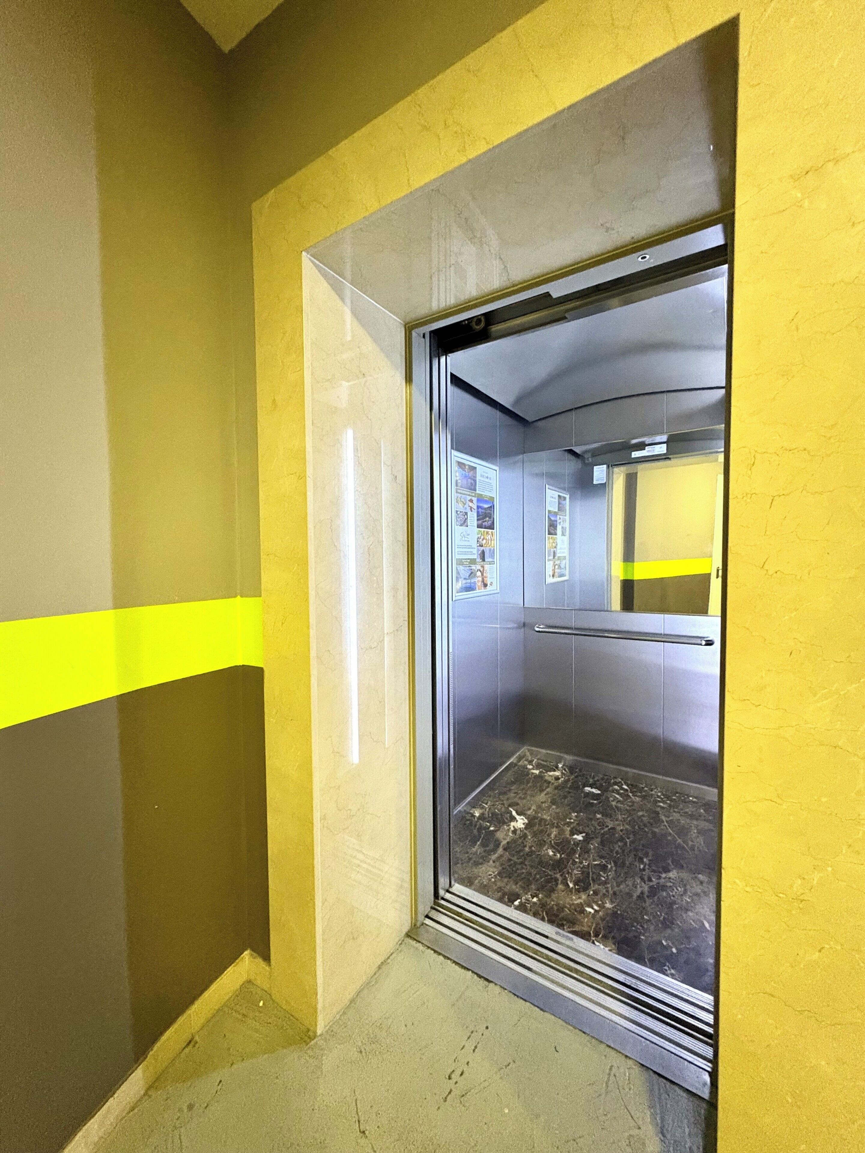 A narrow hallway with yellow and grey walls, a glass elevator door, and a metal handrail.