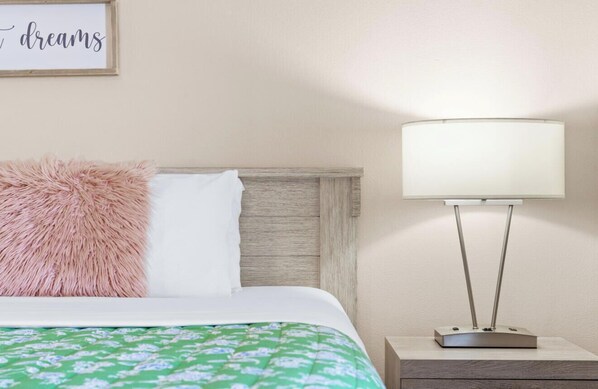 Un lit bien fait, agrémenté d’un oreiller rose moelleux, d’un oreiller blanc et d’une couvre-lit à motifs floraux verts. Un mur blanc est orné d’un cadre contenant une inscription qui dit « rêves ». Une lampe de chevet moderne, avec un abat-jour blanc, est posée sur une table de chevet en bois, à côté du lit.