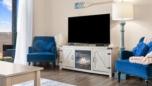 Ein Wohnzimmer mit großem Flachbildfernseher, zwei blauen Sesseln, einem Holzkabinett mit Kamin und Blick ins Freie durch ein großes Fenster.