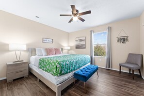 Une chambre à coucher comprenant un lit, des tables de chevet, un ventilateur de plafond, une fenêtre avec des rideaux et une chaise.
