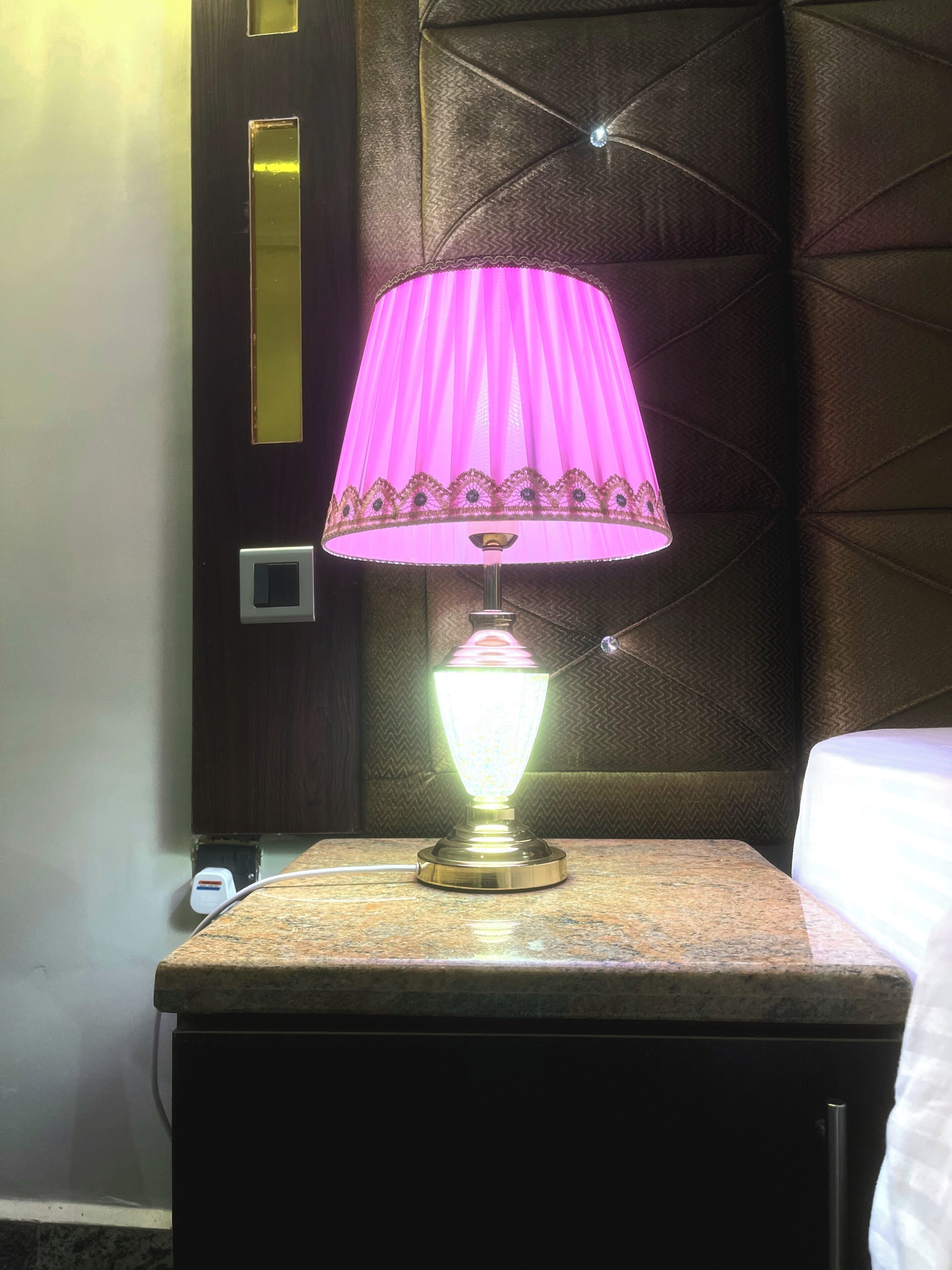 A hotel room with a marble nightstand, a pink lamp, and a dark wooden headboard with a geometric pattern.