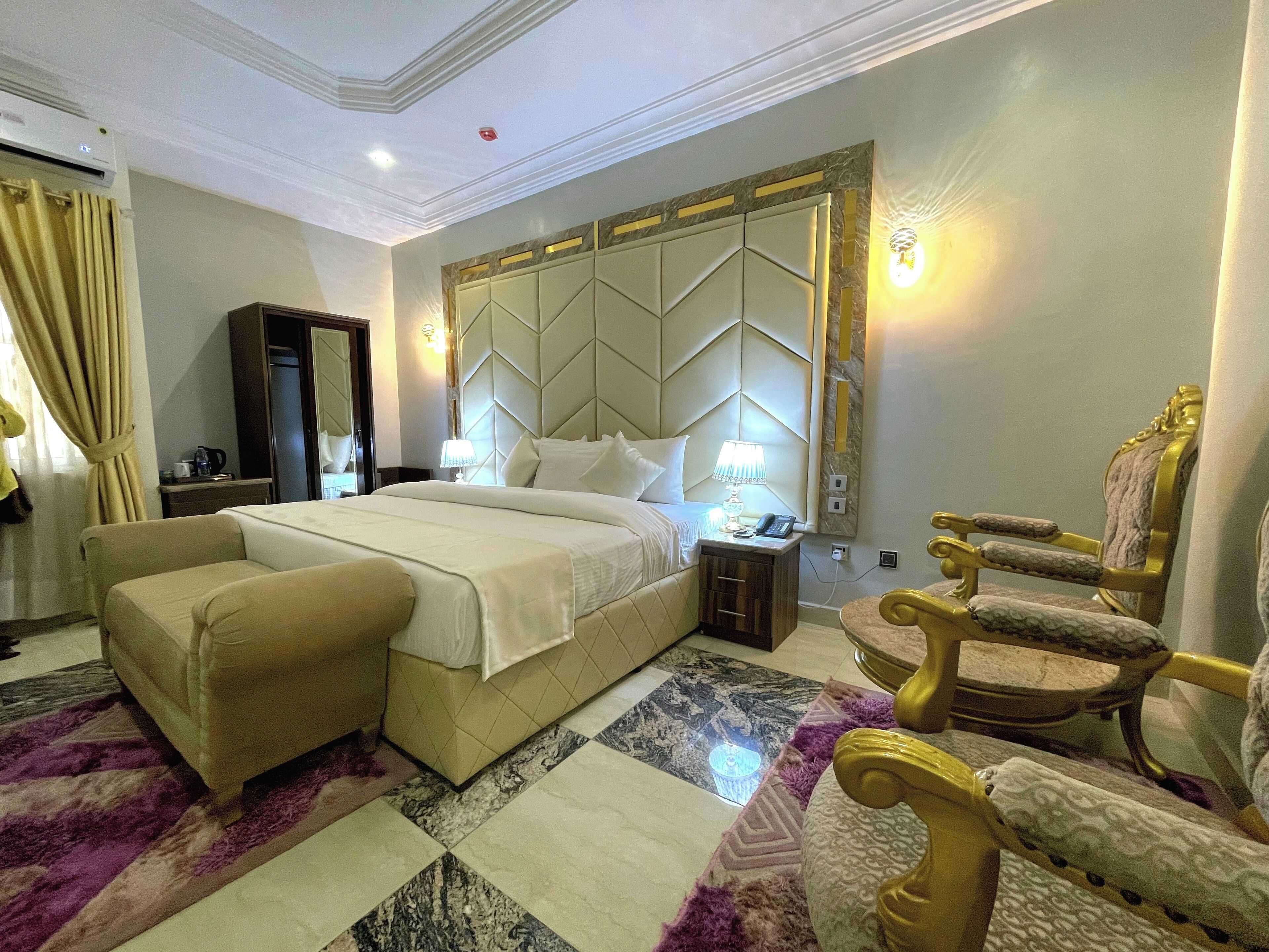 A spacious hotel room with a large bed, a separate sofa, a gold armchair, a marble-patterned floor, and a modern headboard.