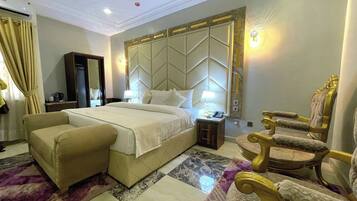 A spacious hotel room with a large bed, a separate sofa, a gold armchair, a marble-patterned floor, and a modern headboard.