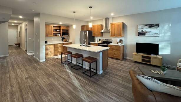 A modern kitchen with wooden cabinets, a central island with stools, and a built-in oven. - SPNC Unit 101 (Fargo)