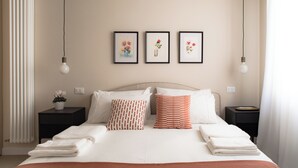 Ein ordentlich gemachtes Bett mit weißen und orangefarbenen Kissen, ein Nachttisch mit Pflanze und drei gerahmte Bilder an der Wand.