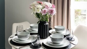 Una mesa de comedor dispuesta con platos, cuencos y vasos blancos, junto a un florero negro con flores.