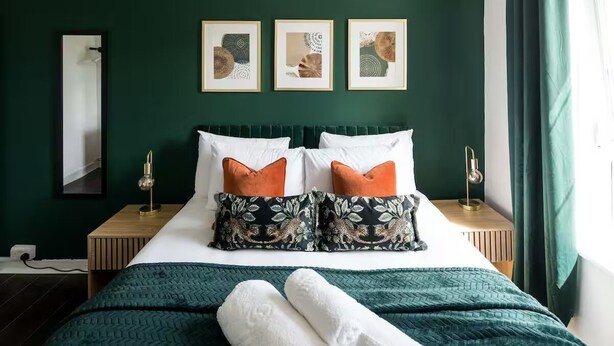 Una cama bien hecha con colcha verde, almohadas blancas y dos almohadas decorativas anaranjadas. Encima de la cama, hay tres cuadros enmarcados en la pared.