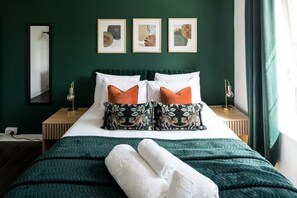 Een netjes opgemaakt bed met een groene sprei, witte kussens en twee oranje decoratieve kussens. Daarboven hangen drie ingelijste foto’s aan de muur.
