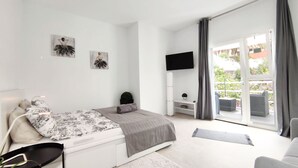 Quarto moderno com cama, TV, uma pequena planta e varanda com cortinas.