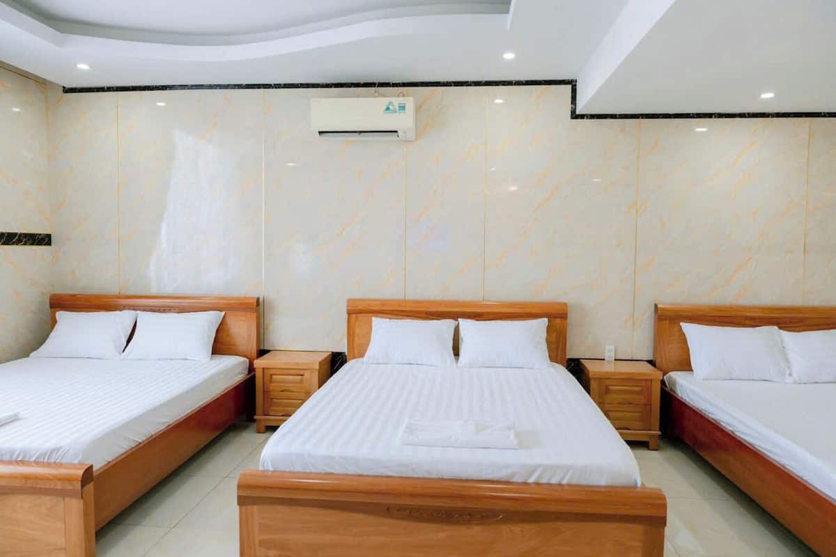 Une chambre d’hôtel avec trois lits en bois, chacun doté d’une tête de lit en bois et de draps blancs. Un appareil de climatisation est fixé au plafond, et une petite table de chevet en bois, avec des tiroirs, se trouve à côté de chaque lit.