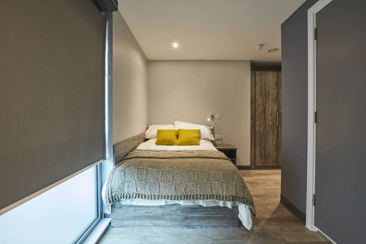 Un corridoio stretto e ben illuminato che conduce a un letto con una coperta a motivi e due cuscini, uno giallo.