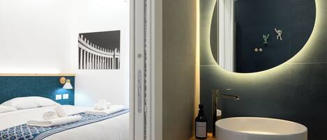 Ein modernes Hotelzimmer mit einem ordentlich bezogenen Bett, einem runden Spiegel mit integriertem Lichtring, einem Holznachttisch mit weißem Waschbecken und einem gerahmten Bild an der Wand.