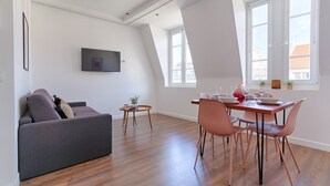 Een moderne woonkamer met een grijze bank, een eettafel met roze stoelen en een flatscreen televisie aan de muur.