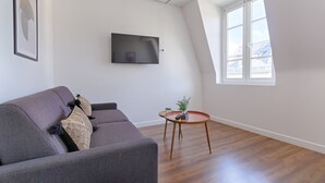 Een moderne woonkamer met een grijze bank, een flatscreen televisie aan de muur, een klein rond tafeltje met een plant, en een groot raam dat natuurlijk licht biedt.