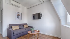 Een moderne woonkamer met een grijze bank, een klein rond tafeltje en een aan de muur bevestigde televisie.