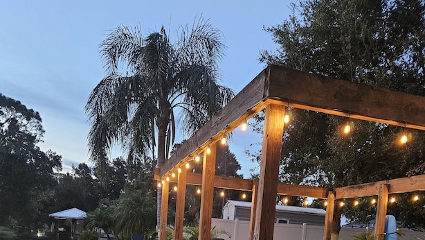 A wooden deck with benches and a pergola with string lights, overlooking a sandy area with a small pool and lounge chairs. - Enjoy Pool, Billiards, Sauna, Access to the Lake (Lake Placid)