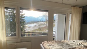 A bedroom with a bed, a bench, a radiator, and a view of a snowy landscape and mountains through large windows. - "Bergnest", Oberstaufen/Steibis (Oberstaufen)