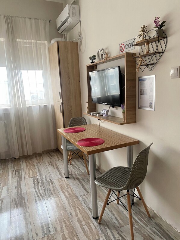Una stanza con un tavolo di legno, due sedie, una TV montata a parete, un piccolo ripiano con oggetti decorativi e un cartello ‘No Smoking’.
