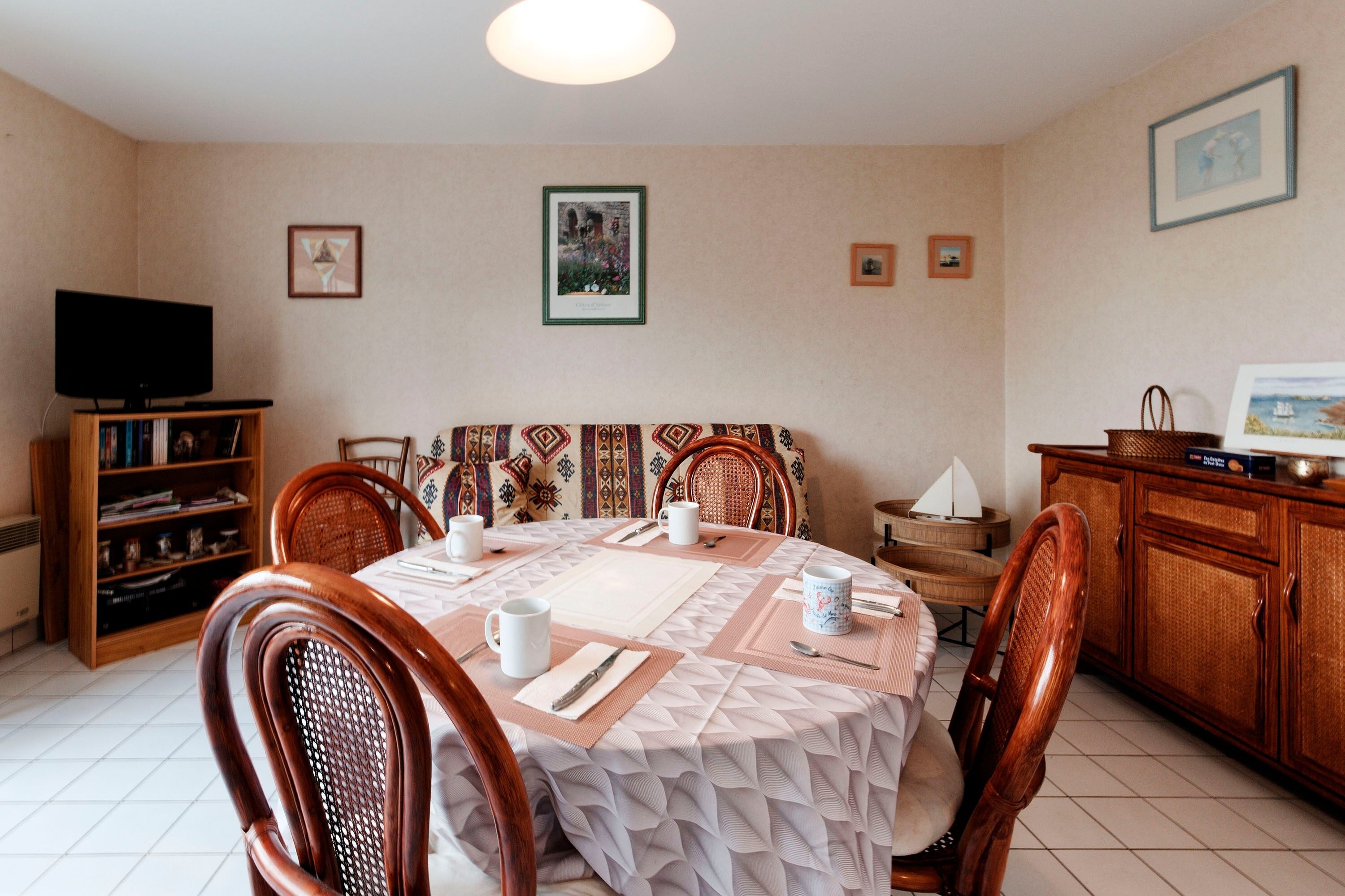 Un comedor con una mesa redonda dispuesta para cuatro personas, rodeada de sillas de mimbre. Un televisor está montado en la pared a la izquierda, y se puede ver una estantería en la esquina.
