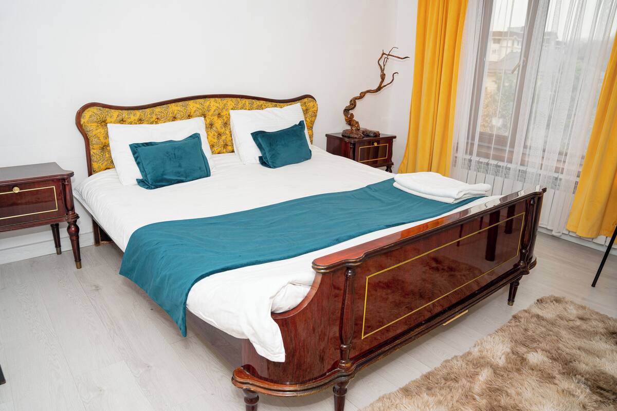 A neatly made bed with a wooden frame and a blue bedspread, a nightstand with a lamp, and a view of the outdoors through a window with yellow curtains.