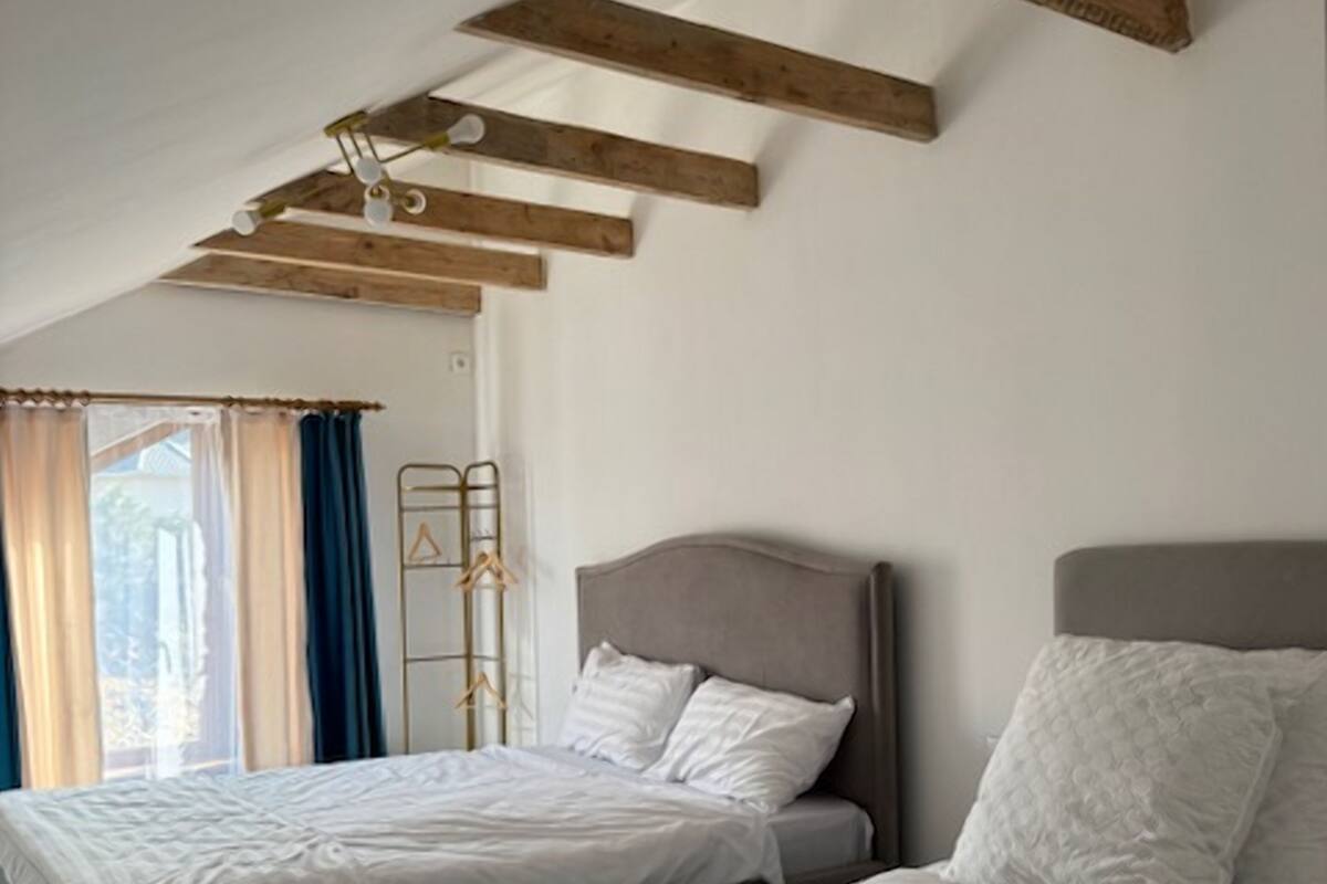 A bedroom with two beds, wooden beams on the ceiling, a window with curtains, and a metal coat rack.
