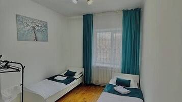 Ein kleiner, gut beleuchteter Raum mit zwei Einzelbetten, einem Deckenventilator, einem Heizkörper und einem fenster mit Vorhängen.