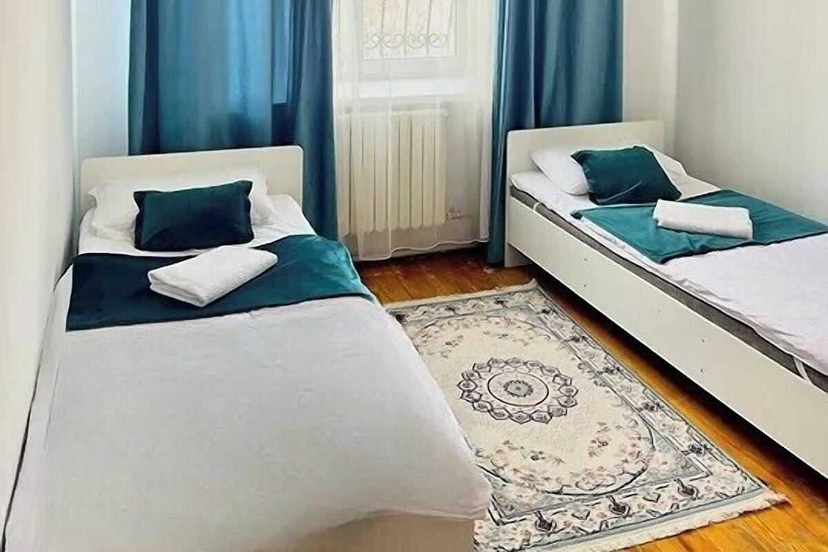 A bedroom with two single beds, a window with curtains, a radiator, and a patterned rug.
