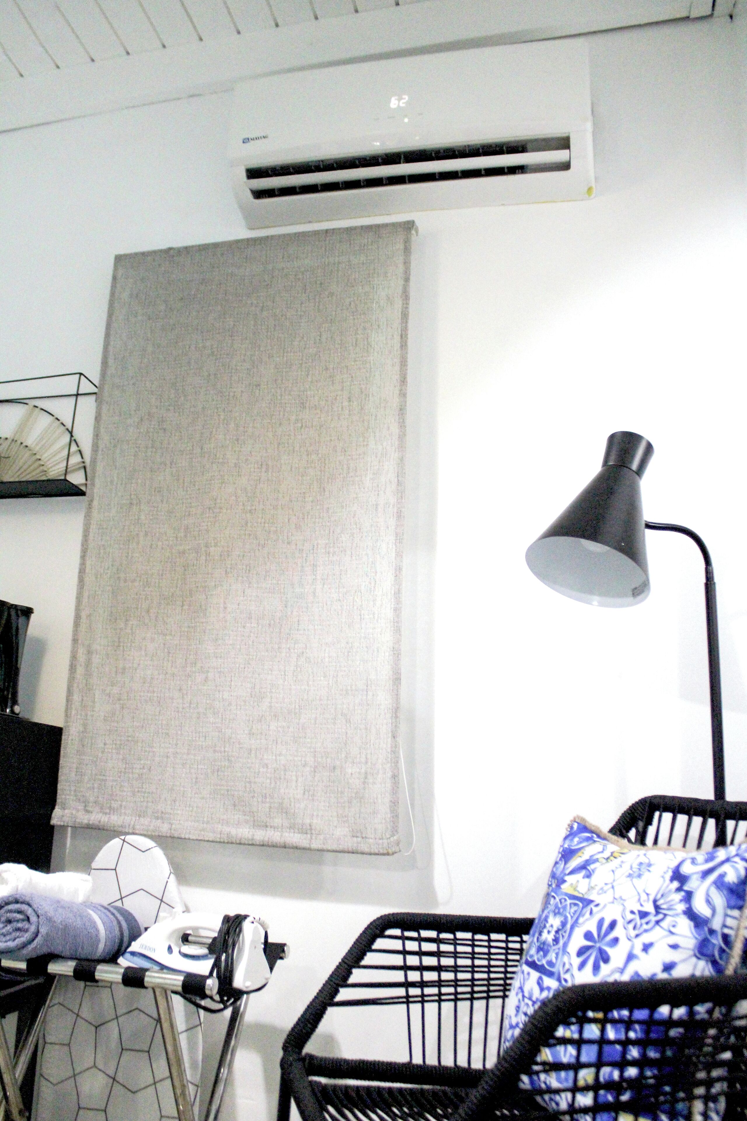 Quarto com uma grande janela coberta por uma cortina bege, um abajur preto, uma cadeira preta com almofada estampada em azul e branco, e um pequeno centro de passar roupa com ferro e toalha enrolada.
