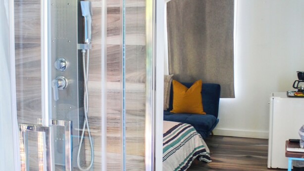 A modern bathroom with a glass-enclosed shower, wooden flooring, a bed with a striped bedspread, and a small table with a coffee maker. - Parguera Beach Club Hotel (Lajas)