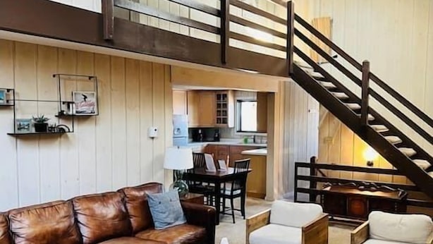 A spacious living room with a brown leather sofa, white armchairs, and a wooden coffee table. A staircase leads to an upper level with a balcony. - 6BR 3BA - MT SNOW, SKIBACK TRAIL, FREE SHUTTLE, POOL, BBQ - Green Mountain House (Dover)