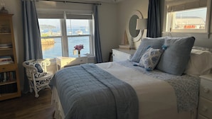 Un dormitorio con una cama grande, una mesita de noche con lámpara, una silla, una estantería y vista al agua.