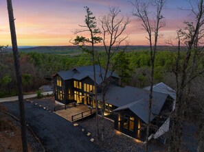 A modern two-story house with large windows and a covered patio area, surrounded by trees and a scenic view. - 5BR Luxury Midnight Skies | 4 King Suites & Hot Tub (Broken Bow)