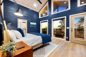 A bedroom with a bed, bedside table, lamp, and a view of the outdoors through a sliding door. - 5BR Luxury Midnight Skies | 4 King Suites & Hot Tub (Broken Bow)
