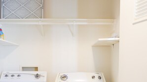 Une buanderie équipée de deux machines à laver blanches, d’une bouche d’aération et d’une étagère.