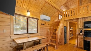Interior de una cabaña de madera con una mesa de comedor, banco, escalera y zona de cocina.