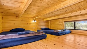Una cabaña con paredes y suelos de madera, dos camas con ropa de cama azul, una ventana que da a árboles y un techo con vigas de madera.