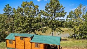 Una cabaña de madera con techo verde, ubicada en una zona arenosa con un lago y árboles al fondo.