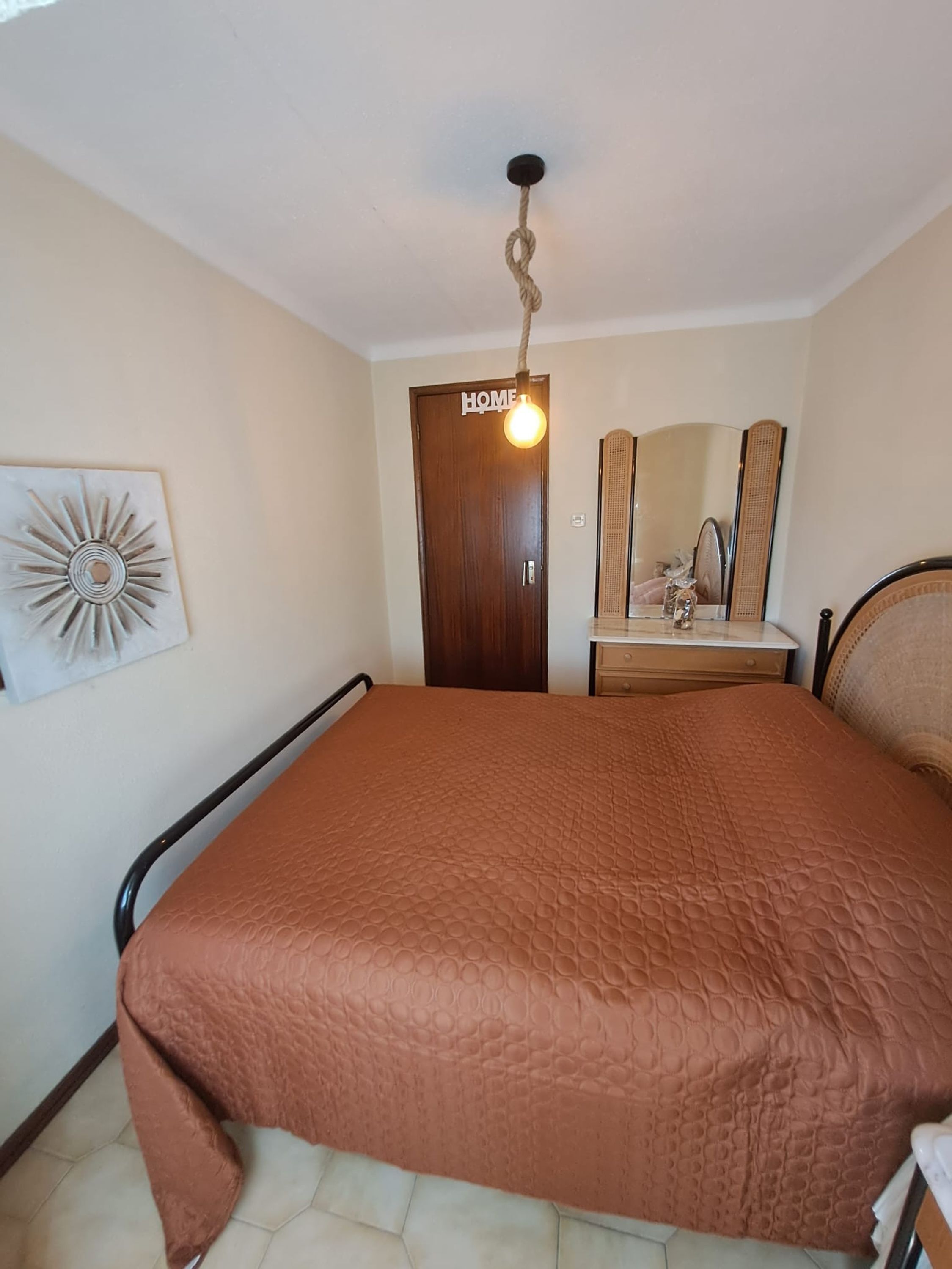 A bedroom with a bed, a wooden door, a mirror, and a wall-mounted light fixture.