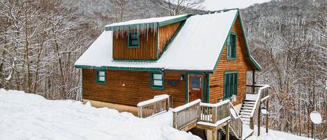 A wooden cabin with a steeply pitched roof, a front porch, and a staircase leading to a deck.
