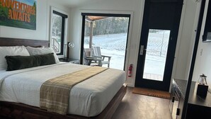 A bedroom with a large bed, a wooden headboard, a nightstand, and a desk with a chair. There is a door leading to a balcony with a view of a snowy landscape.