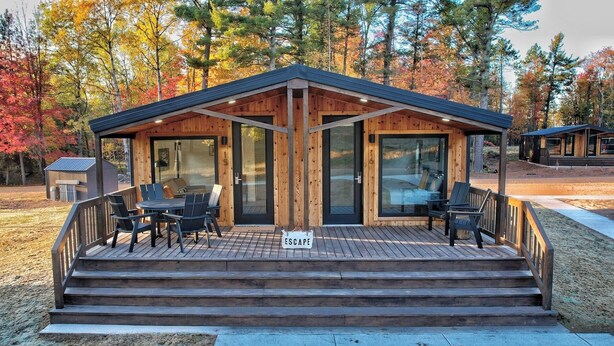 A wooden cabin with a deck, furnished with chairs and a table, surrounded by autumn trees.