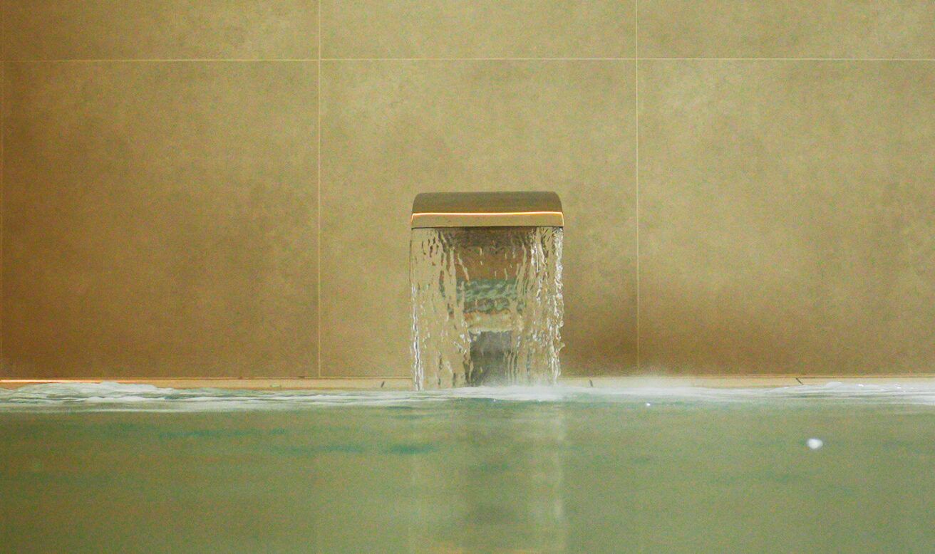Ein Wasserbecken mit quadratischer Basis, aus dem Wasser fließt, vor einer schlichten Wand.