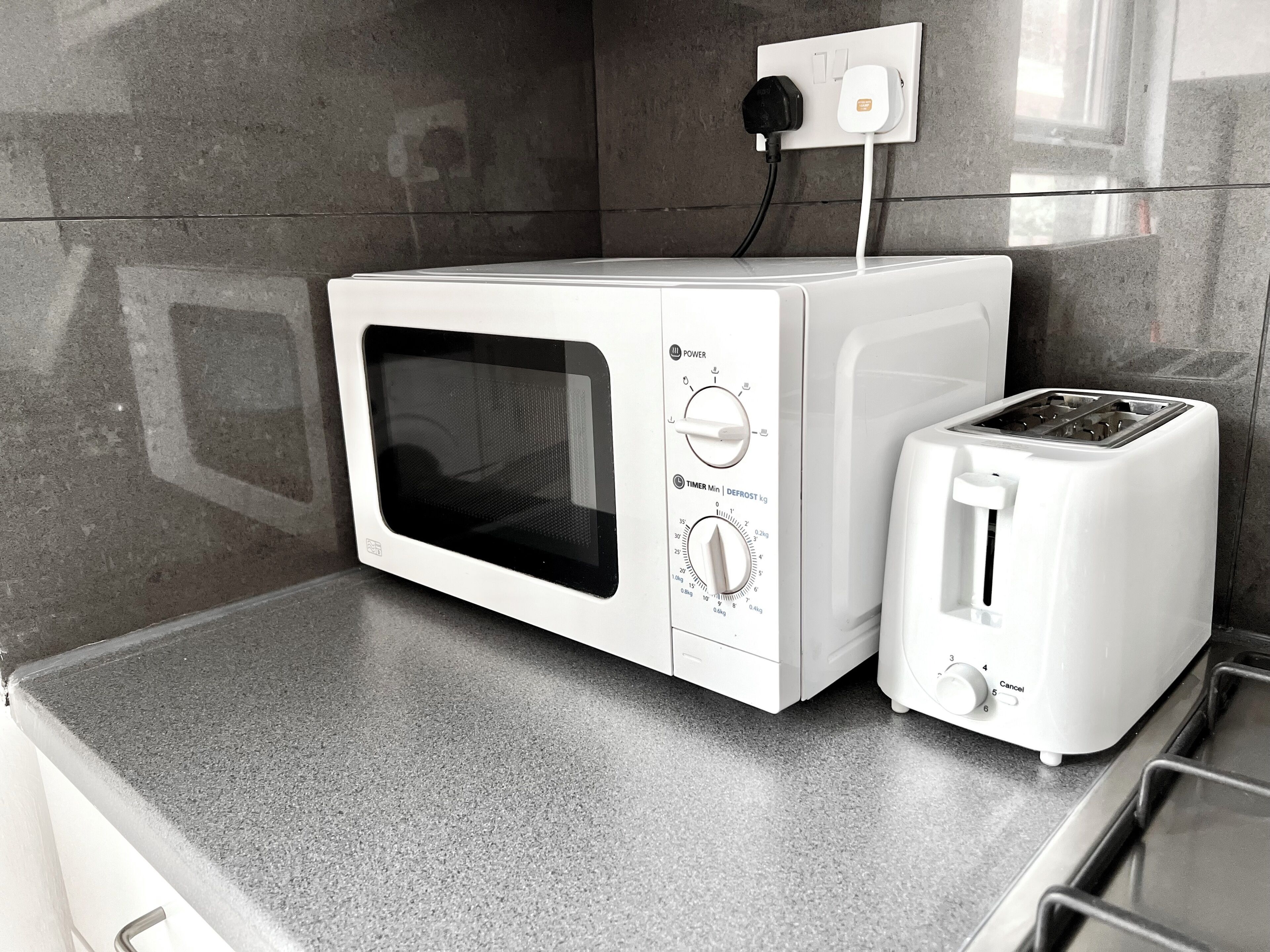 A kitchen with a white microwave and toaster on a grey countertop, a power outlet above, and a tiled backsplash.