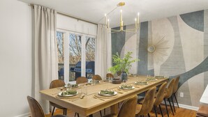 A dining area with a long table set for six, surrounded by chairs, a chandelier, and a large window with curtains.