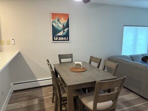 Een eethoek met een houten tafel en stoelen, een poster aan de muur, en een raam met jaloezieën.