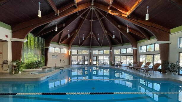 An indoor swimming pool with a high ceiling and wooden beams, surrounded by windows and lounge chairs.