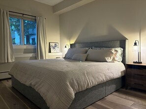 Een slaapkamer met een groot bed, een nachtkastje, een lamp en een raam met gordijnen.