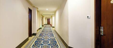 A hotel hallway with a patterned carpet, a wooden door on the right, and a light fixture on the ceiling.