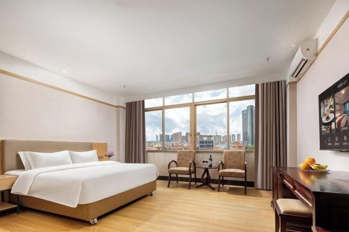 A hotel room with a large bed, two chairs, a small table, and a city view through large windows.